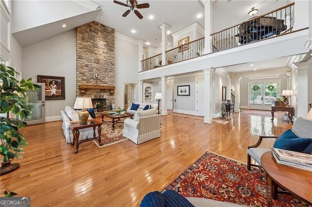living room featuring a ceiling fan, a stone fireplace, light wood finished floors, decorative columns, and high vaulted ceiling