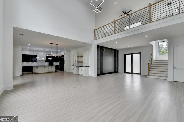 unfurnished living room featuring light wood-style flooring, a high ceiling, baseboards, ceiling fan, and stairs