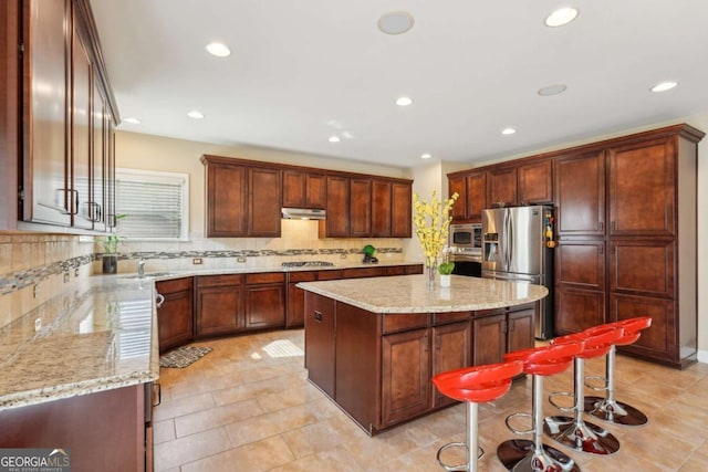 kitchen featuring under cabinet range hood, decorative backsplash, a center island, light stone countertops, and appliances with stainless steel finishes