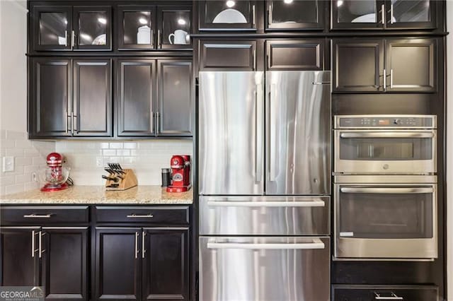 kitchen featuring stainless steel appliances, glass insert cabinets, decorative backsplash, and light stone countertops