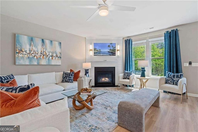 living room featuring a ceiling fan, baseboards, visible vents, wood finished floors, and a large fireplace