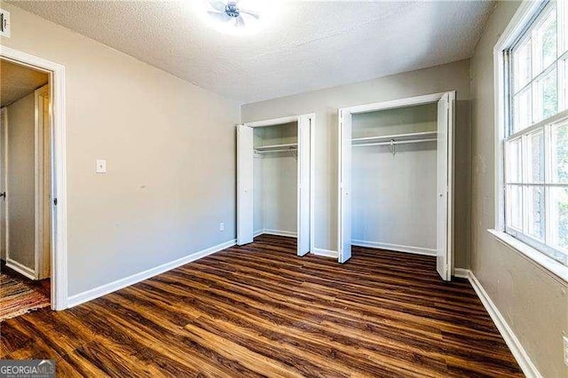unfurnished bedroom featuring a textured ceiling, baseboards, dark wood-style floors, and multiple closets