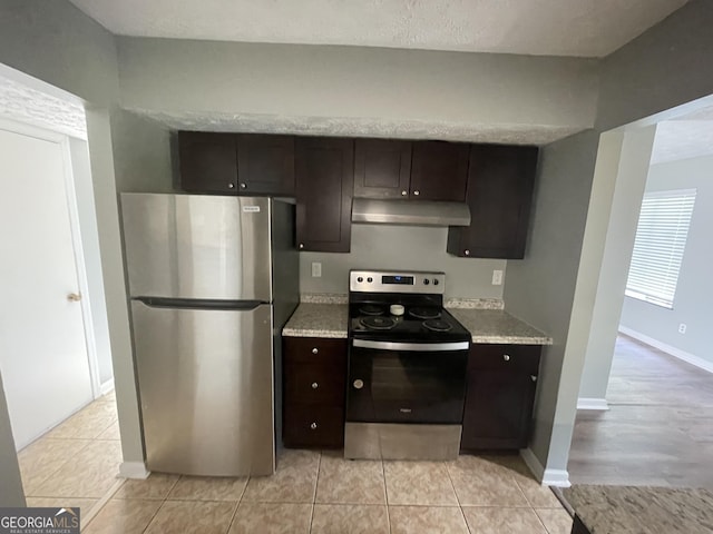kitchen featuring under cabinet range hood, baseboards, light tile patterned flooring, dark brown cabinetry, and stainless steel appliances