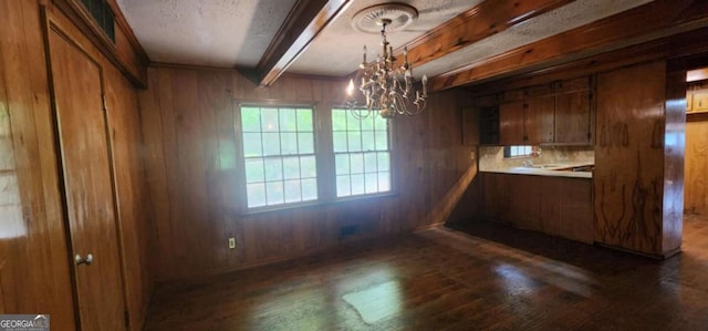 unfurnished dining area featuring wood walls, a textured ceiling, dark wood finished floors, beamed ceiling, and a notable chandelier