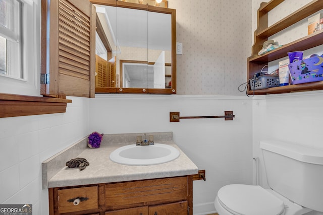 bathroom with tile walls, wallpapered walls, toilet, vanity, and a wainscoted wall