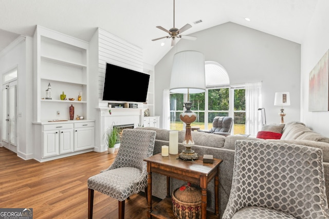 living room featuring light wood-style flooring, a ceiling fan, high vaulted ceiling, a fireplace, and built in shelves