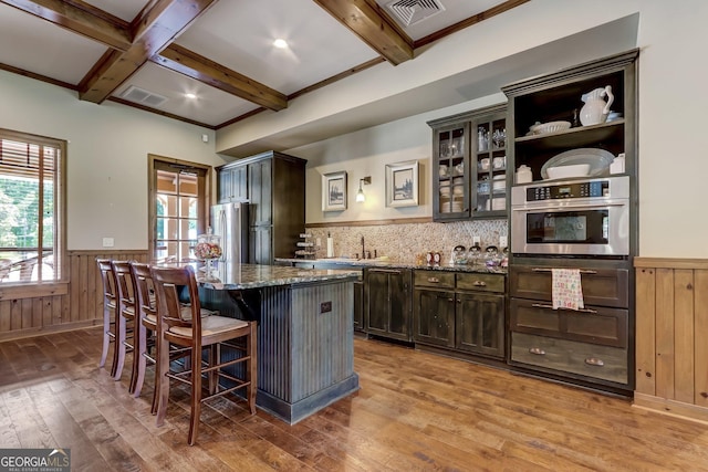 kitchen with wainscoting, beam ceiling, stainless steel appliances, dark stone counters, and light wood finished floors