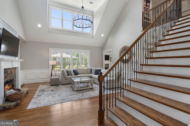 living area with high vaulted ceiling, a stone fireplace, stairs, wood finished floors, and wainscoting