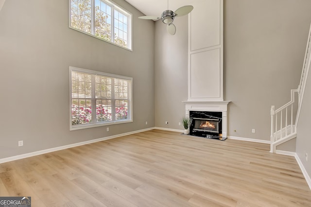 unfurnished living room with a ceiling fan, a fireplace, light wood-style flooring, baseboards, and a high ceiling