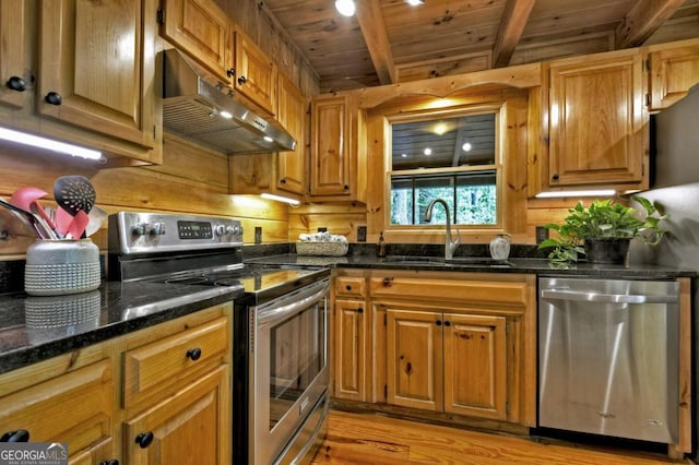 kitchen with a sink, under cabinet range hood, light wood finished floors, beamed ceiling, and appliances with stainless steel finishes