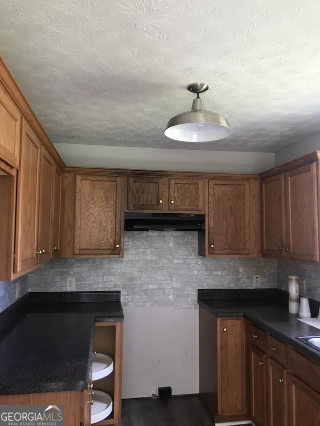 kitchen with a textured ceiling, under cabinet range hood, decorative backsplash, brown cabinets, and dark countertops