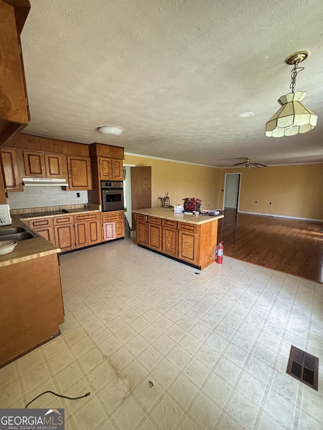 kitchen featuring oven, brown cabinets, a ceiling fan, under cabinet range hood, and a textured ceiling