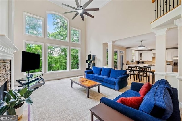 living area with ceiling fan, decorative columns, a stone fireplace, a high ceiling, and light carpet