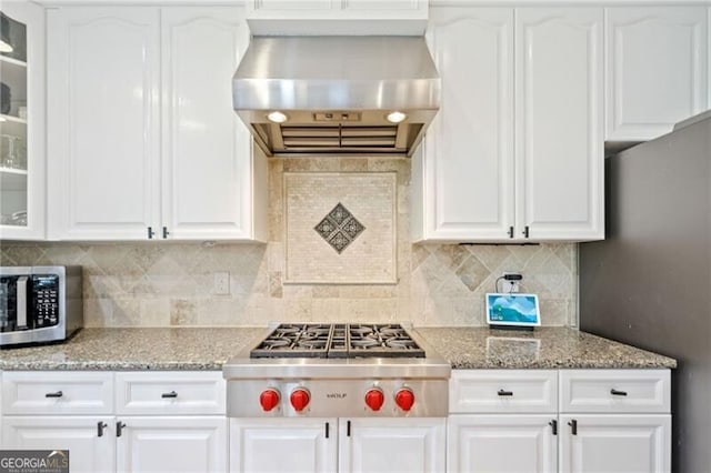 kitchen with wall chimney exhaust hood, appliances with stainless steel finishes, glass insert cabinets, white cabinets, and light stone counters