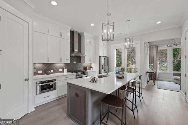 kitchen featuring stainless steel appliances, wall chimney range hood, tasteful backsplash, a kitchen island with sink, and an inviting chandelier