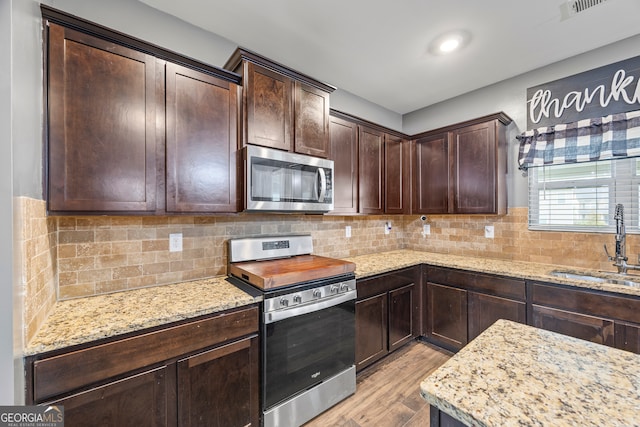 kitchen with appliances with stainless steel finishes, a sink, light wood-type flooring, dark brown cabinetry, and light stone counters