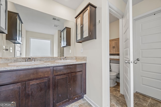 bathroom featuring stone tile flooring, toilet, double vanity, and baseboards