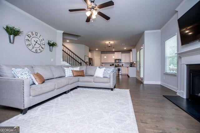 living area with baseboards, dark wood-style flooring, a glass covered fireplace, ceiling fan, and crown molding
