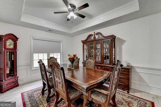 dining space with a ceiling fan, light wood finished floors, a decorative wall, crown molding, and a raised ceiling