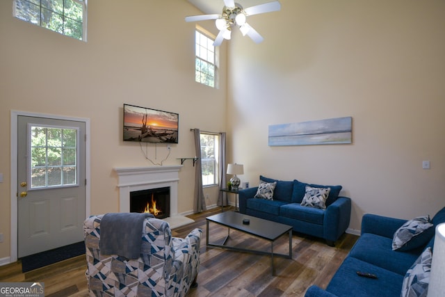 living area with wood finished floors, a warm lit fireplace, a towering ceiling, a ceiling fan, and baseboards