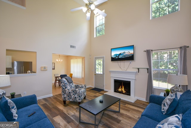 living room featuring a towering ceiling, a chandelier, wood finished floors, a warm lit fireplace, and ceiling fan
