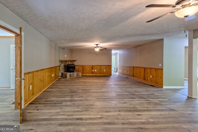 unfurnished living room with a ceiling fan, a wainscoted wall, wooden walls, wood finished floors, and a stone fireplace