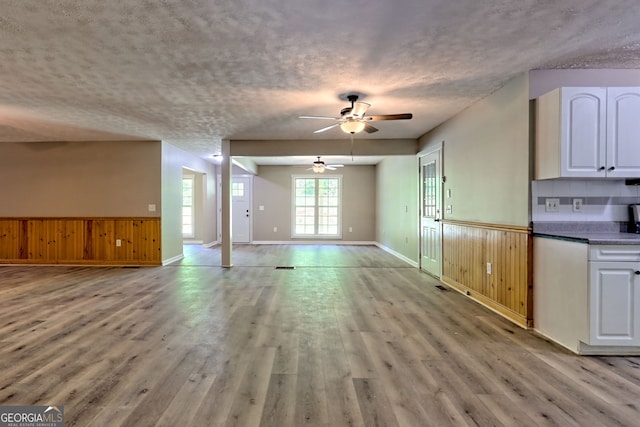 unfurnished living room featuring light wood-type flooring, ceiling fan, a textured ceiling, a wainscoted wall, and wooden walls