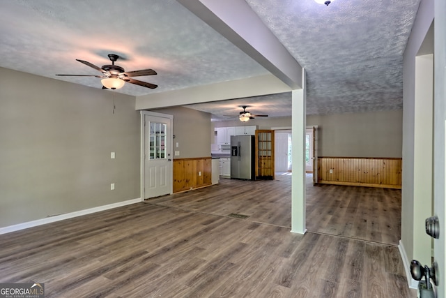 unfurnished living room featuring a ceiling fan, dark wood-style floors, wainscoting, baseboards, and wood walls