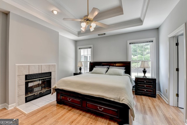 bedroom featuring light wood-style flooring, ornamental molding, a tile fireplace, a raised ceiling, and baseboards