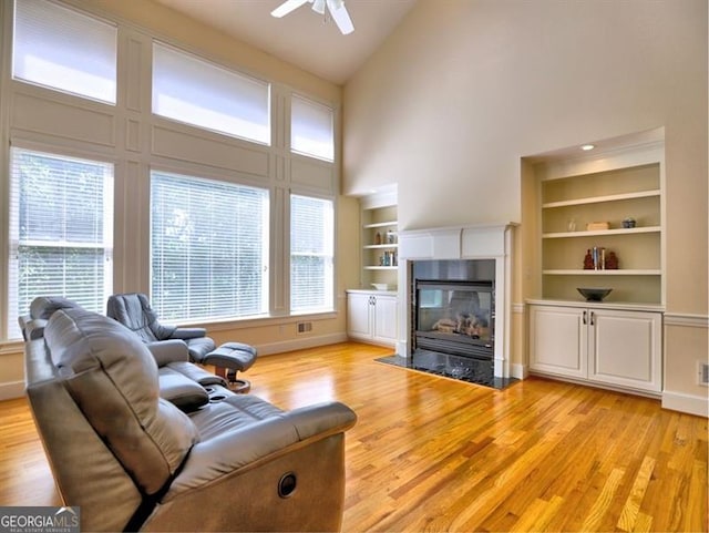 living area featuring high vaulted ceiling, built in features, light wood-style flooring, a fireplace with flush hearth, and ceiling fan