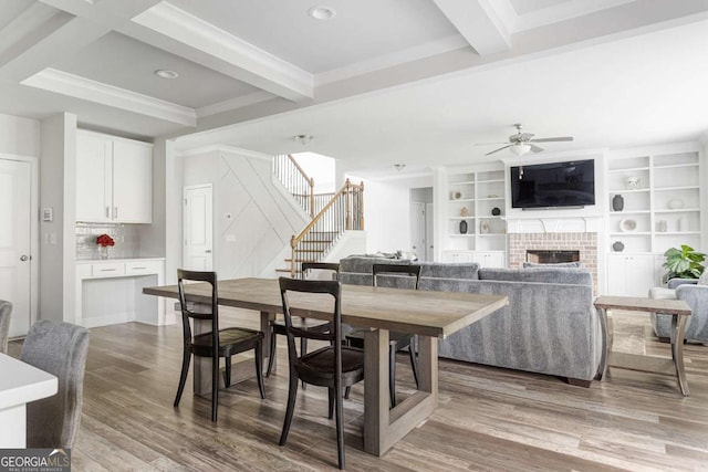dining room with ceiling fan, beamed ceiling, stairs, wood finished floors, and a brick fireplace