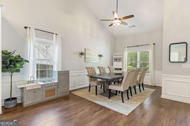 dining room featuring high vaulted ceiling, a decorative wall, a ceiling fan, dark wood-type flooring, and wainscoting