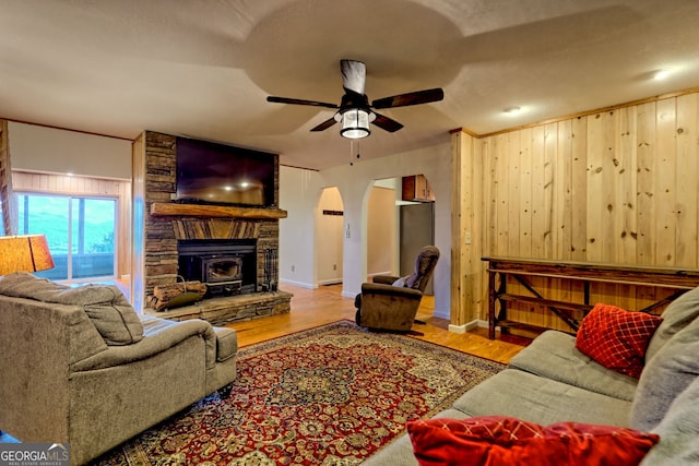 living area featuring wood finished floors, arched walkways, ceiling fan, a stone fireplace, and wooden walls