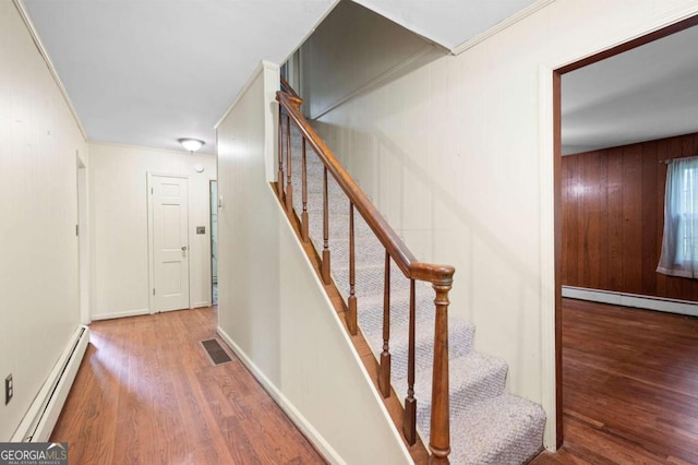 stairway with baseboard heating, wood finished floors, wooden walls, and ornamental molding