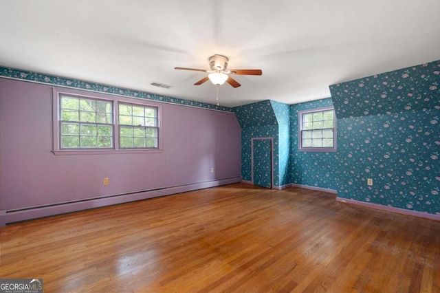 unfurnished room with wallpapered walls, a ceiling fan, wood finished floors, a baseboard heating unit, and baseboards