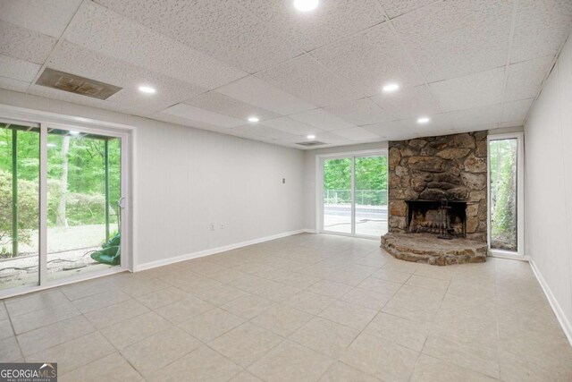 unfurnished living room featuring plenty of natural light, baseboards, a stone fireplace, a drop ceiling, and light tile patterned floors