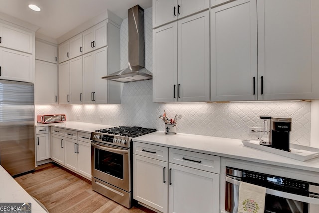 kitchen featuring stainless steel appliances, wall chimney exhaust hood, light countertops, light wood-style floors, and backsplash