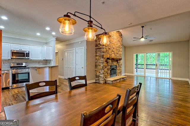dining area featuring ceiling fan, ornamental molding, a stone fireplace, dark wood finished floors, and baseboards