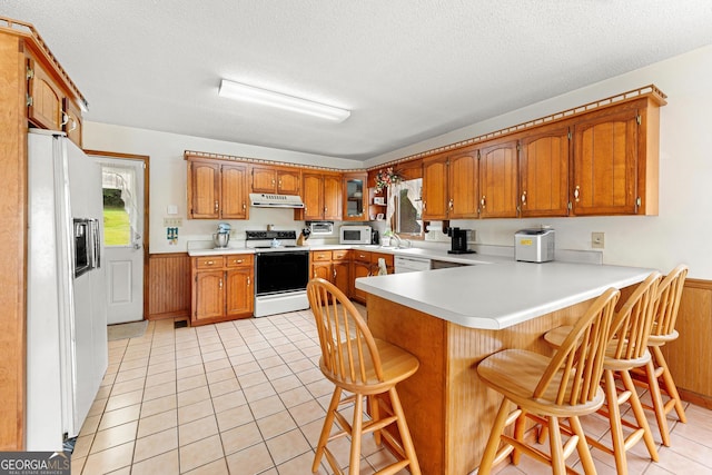 kitchen with white appliances, a peninsula, brown cabinetry, under cabinet range hood, and light countertops