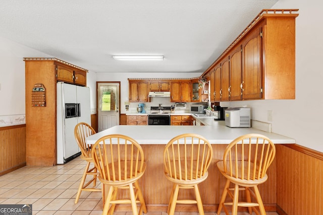 kitchen with white appliances, under cabinet range hood, a peninsula, brown cabinetry, and light countertops