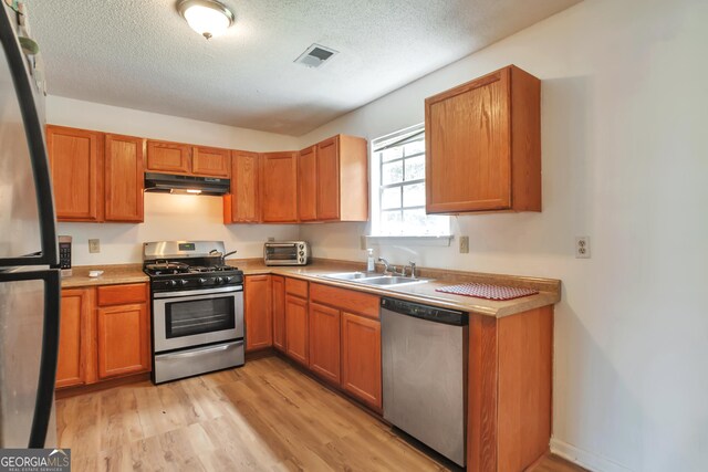 kitchen with stainless steel appliances, under cabinet range hood, a sink, light wood finished floors, and a textured ceiling