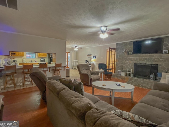 living room with wood finished floors, a ceiling fan, ornamental molding, a textured ceiling, and a stone fireplace