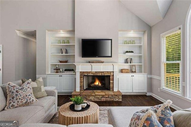 living area with dark wood-style flooring, vaulted ceiling, a stone fireplace, baseboards, and built in shelves