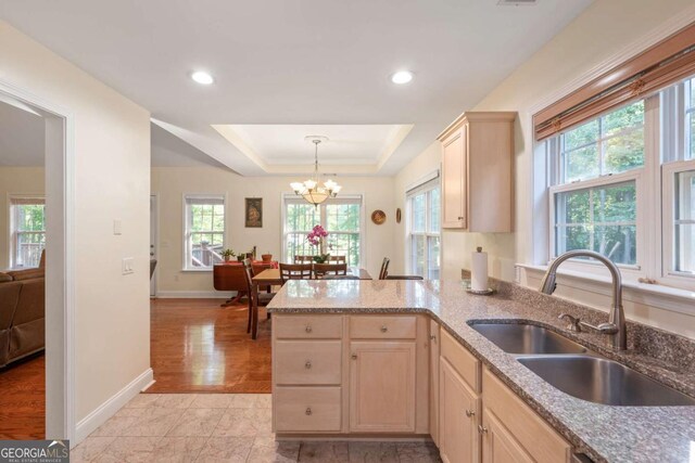 kitchen with a sink, light brown cabinetry, a chandelier, a tray ceiling, and light stone countertops