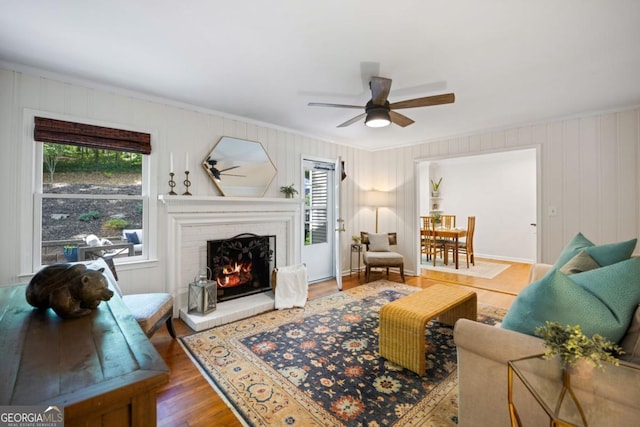 living room featuring crown molding, a ceiling fan, a warm lit fireplace, and wood finished floors
