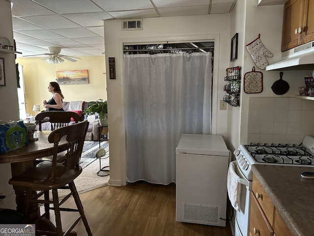 kitchen featuring white gas stove, refrigerator, a drop ceiling, brown cabinets, and under cabinet range hood