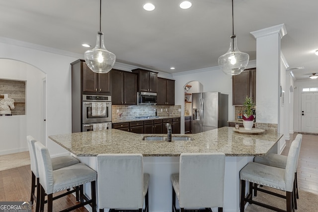 kitchen with stainless steel appliances, arched walkways, a sink, dark brown cabinetry, and light wood-style floors