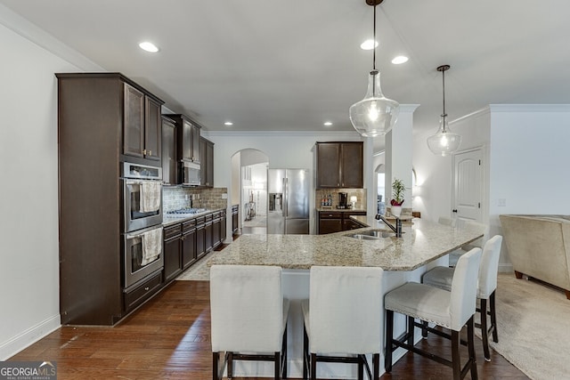 kitchen featuring stainless steel appliances, arched walkways, a sink, dark brown cabinets, and crown molding