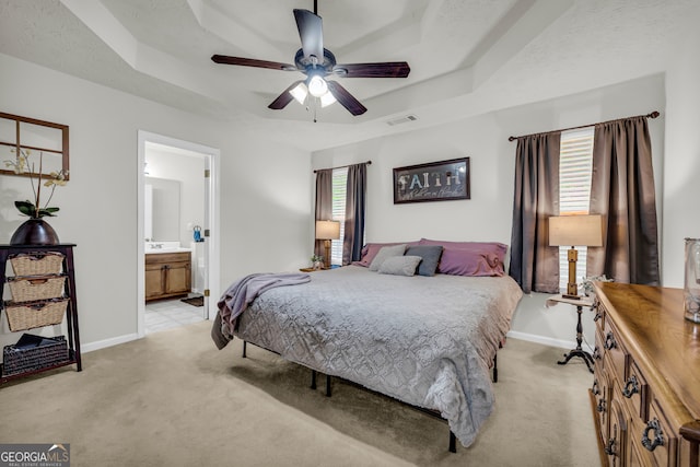 bedroom with a tray ceiling, light colored carpet, baseboards, and ensuite bath