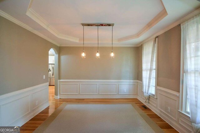 unfurnished dining area with a raised ceiling, wood finished floors, crown molding, wainscoting, and arched walkways